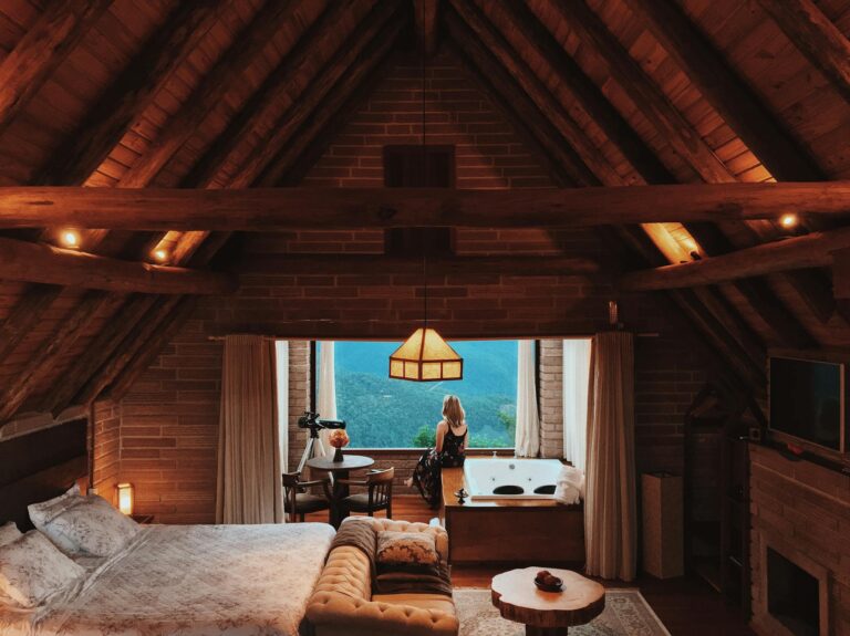 Back view female looking out window while sitting in spacious comfortable mansard room with cozy bed and fireplace in tropical resort